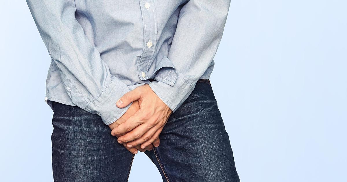 Close up of a man holding his crotch in jeans and a shirt on a light blue background