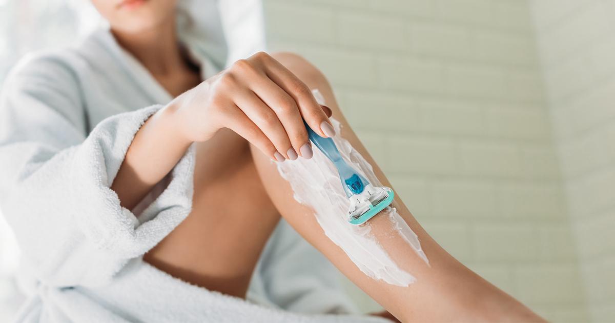 Woman in a bathrobe shaving her leg with a razor.