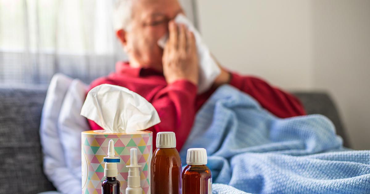 Old man lying on bed blowing his nose with hay fever medicines in the foreground.