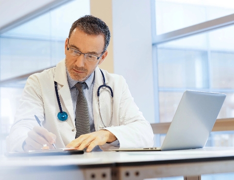 Um médico com óculos e estetoscópio trabalhando em um laptop em uma mesa.