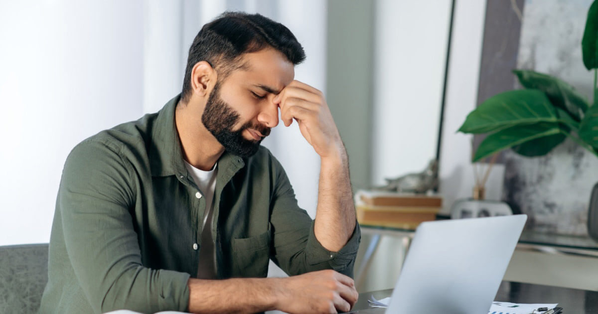Homem sentado à secretária a esfregar os olhos por estar cansado.
